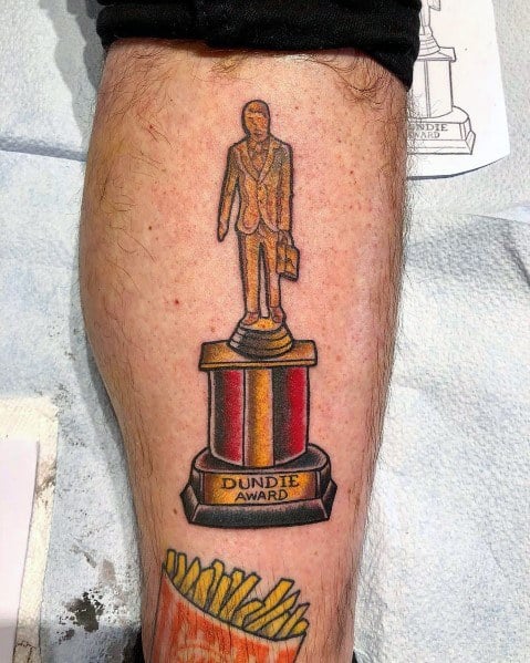 Color tattoo of a Dundie Award trophy from *The Office*, featuring the classic gold figure on a red and yellow pedestal.