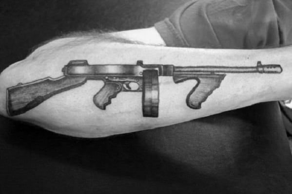 Realistic black and grey Tommy gun tattoo on the forearm, featuring detailed shading and fine linework for a bold and classic firearm design.