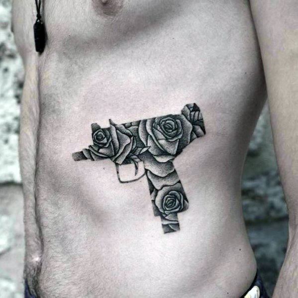 Uzi tattoo on the ribcage with intricate black and grey roses filling the gun’s shape, blending floral elegance with a bold firearm design.