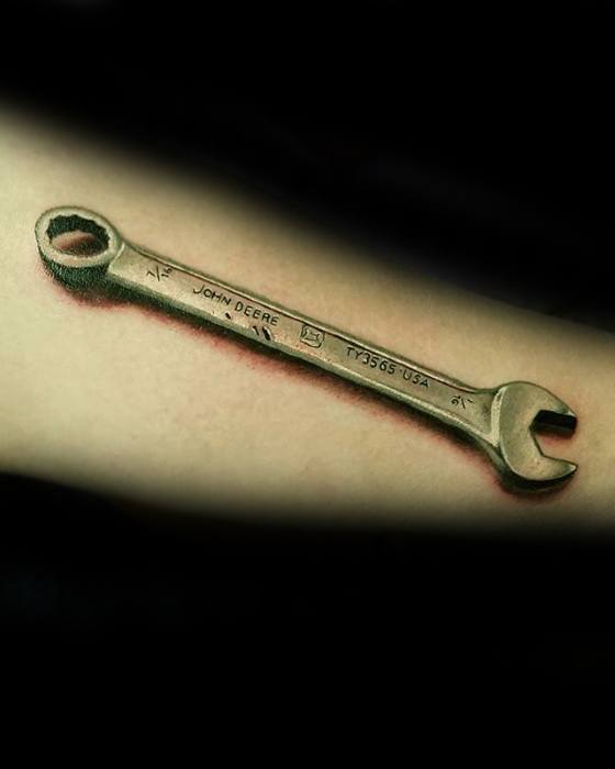 Hyper-realistic tattoo of a John Deere wrench with engraved text and shadowing, creating a 3D effect on the forearm.