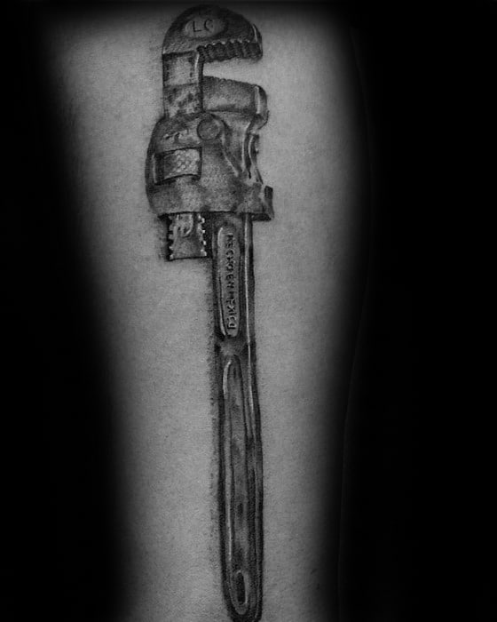 Black and grey realistic tattoo of a pipe wrench with detailed texture and engraved markings on the handle.