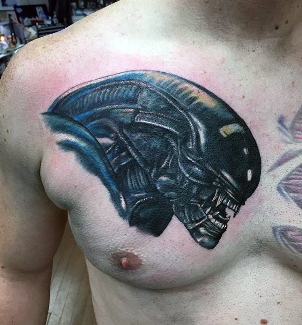 Realistic Xenomorph head tattoo on the chest with metallic sheen, sharp teeth, and bold black and blue shading.