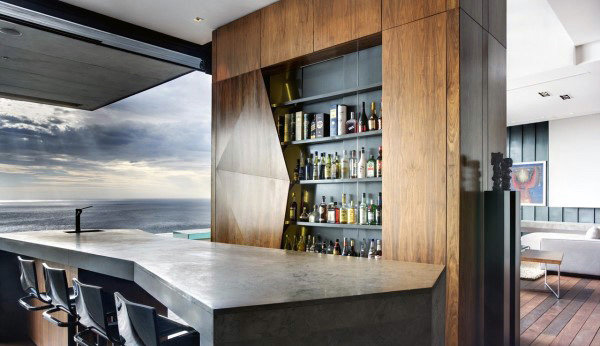 Modern home bar with a sleek countertop, wooden cabinet, and bar stools overlooking the ocean through large windows