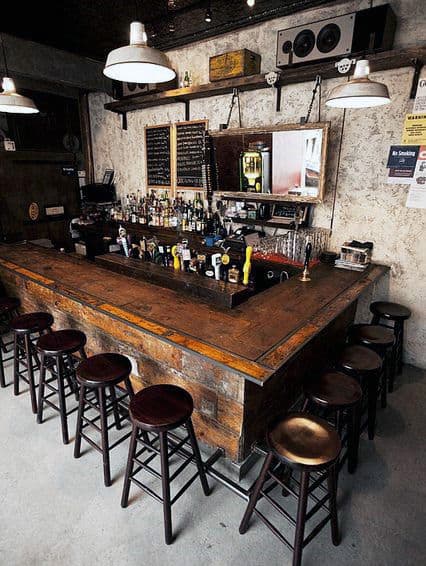 Rustic bar with wooden counter, barstools, and various bottles; warm lighting and chalkboard menu adorn the cozy bar