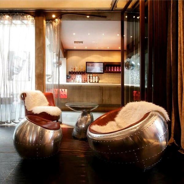 Modern lounge with metallic chairs, fur cushions, glass table, and a bar with soft lighting