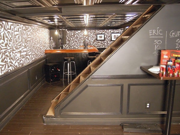 A modern, stylish basement bar with geometric wallpaper and seating under a staircase