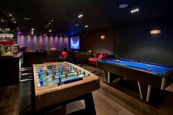 Game room with foosball, pool table, bar, and a screen on the wall, dimly lit with colorful lights