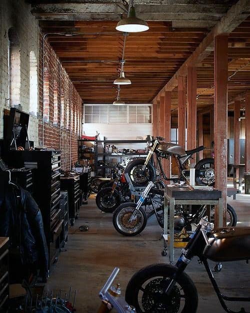 A rustic garage workshop with motorcycles, toolboxes, and wooden beams, illuminated by overhead lights