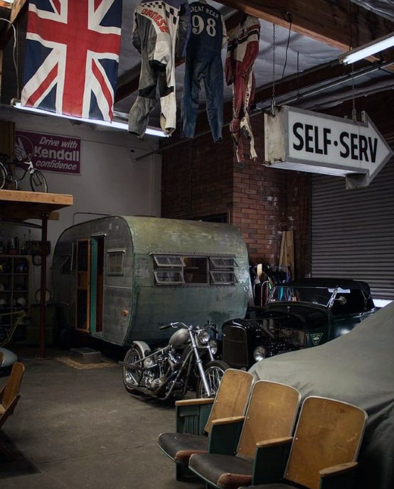 A vintage garage with a Union Jack flag, motorcycle, car, camper, and "Self-Serv" sign hanging from the ceiling