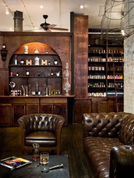 Cozy lounge with leather chairs, a wooden bar, shelves of bottles, and warm lighting