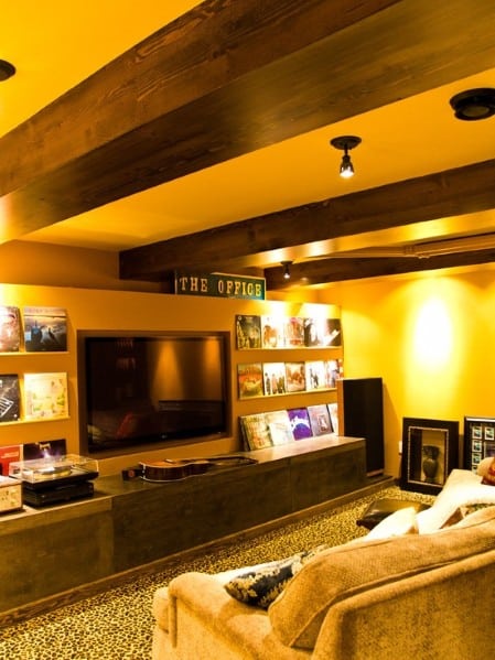 Cozy basement lounge with wall-mounted TV, vinyl records, and a guitar; warm lighting and plush seating add comfort