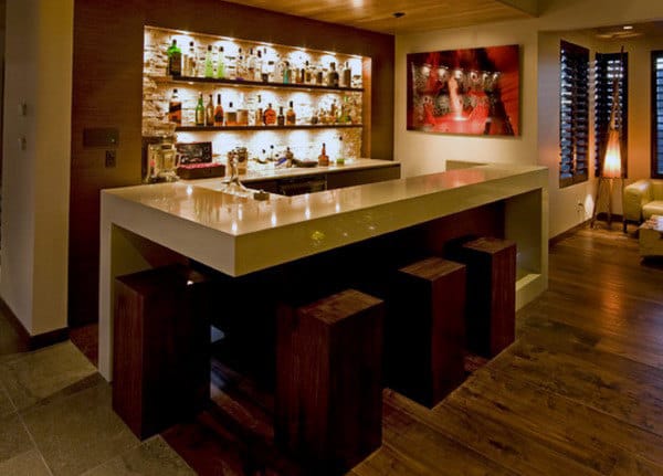 Modern bar with a sleek white countertop, illuminated shelves displaying bottles, and wooden stools