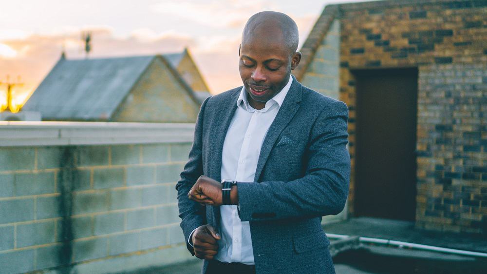 man on a rooftop checks his watch, wearing a grey blazer and white button up