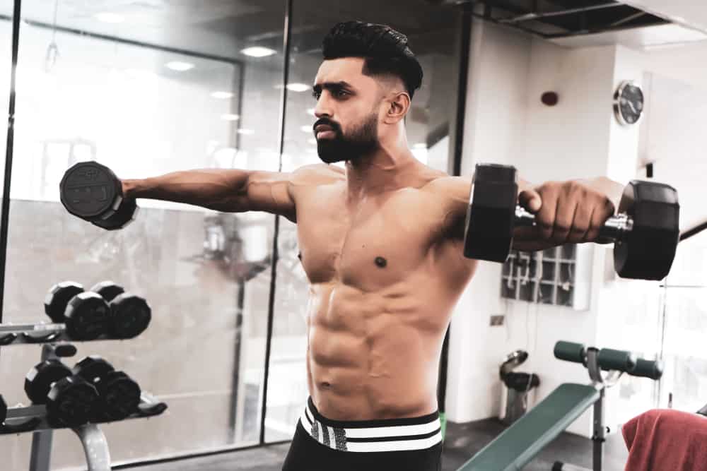 shirtless young man doing side lateral rises exercise in gym