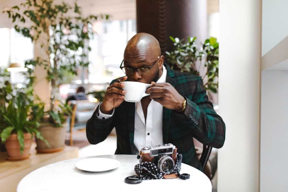 man sits in a cafe, drinking a coffee, wearing a green plaid blazer, and white button up