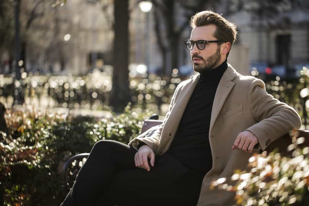 man sits on a park bench in black jeans, a black turtleneck, fitted tan trench coat, and back sunglasses