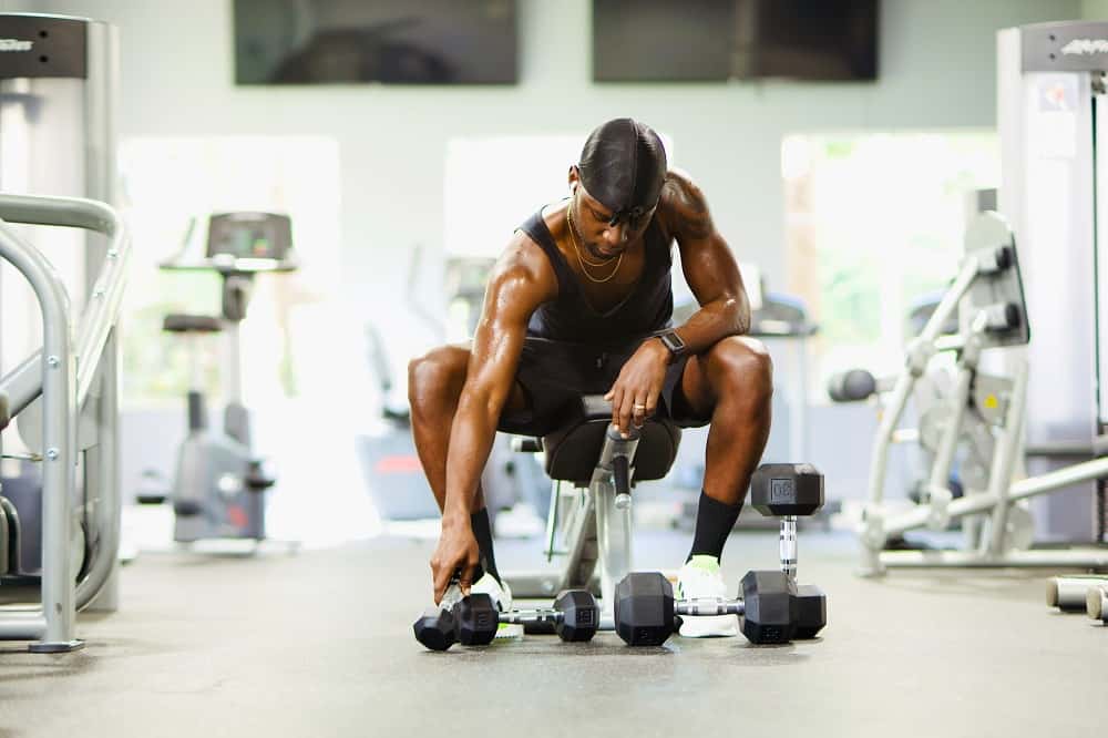 man in a gym sits on a workout bench, looking at various sizes of dumbbells on the ground below him