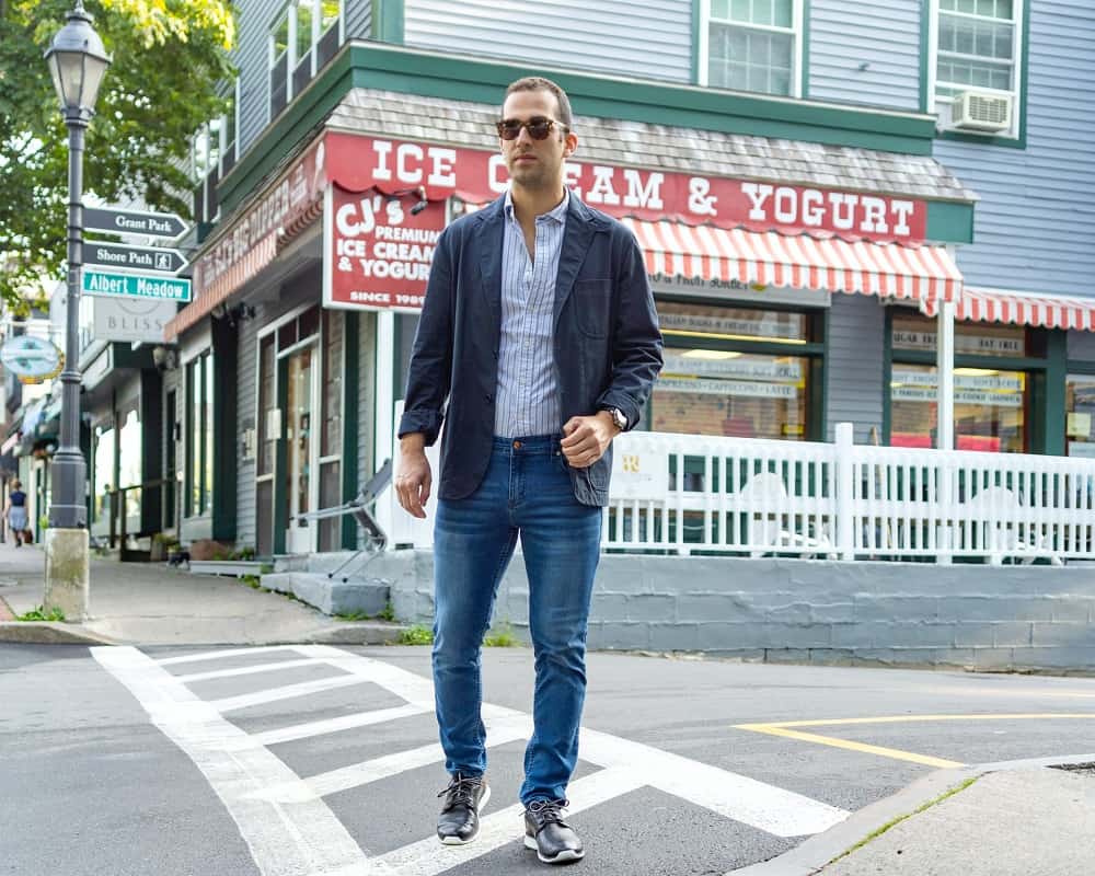 man crosses the street, wearing straight leg denim pants, a collared dress shirt, blazer, and sneakers