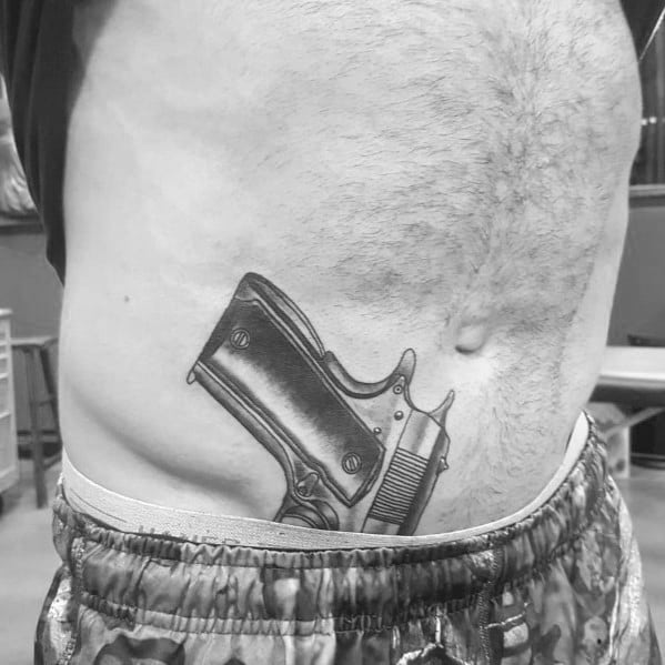 Realistic black and gray tattoo of a 1911 pistol on the lower abdomen, designed to appear as if it is tucked into the waistband for a bold effect.
