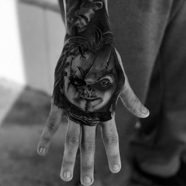 Black and grey Chucky hand tattoo with intense shading, featuring his stitched face, eerie grin, and piercing gaze for a bold horror-inspired design.