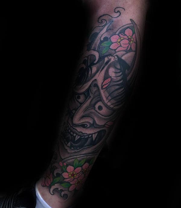 Forearm tattoo featuring a fierce Hannya mask with intricate shading, surrounded by delicate pink cherry blossoms and swirling wind patterns
