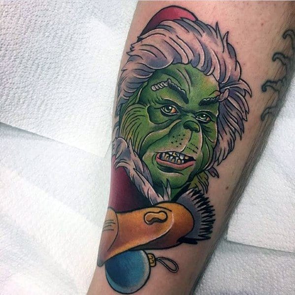 Christmas tattoo of the Grinch wearing a Santa hat, smirking mischievously, surrounded by festive elements like a broken ornament and holiday decorations.