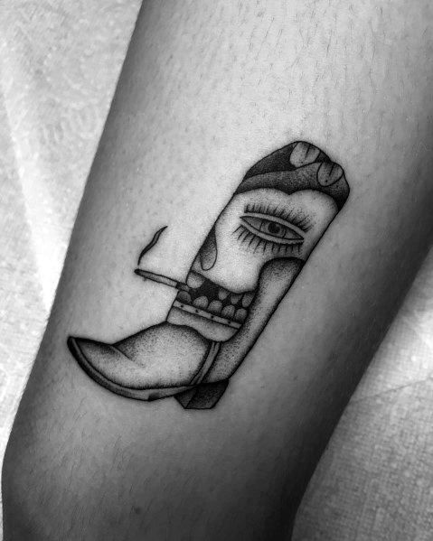 Black and white tattoo of a cowboy boot creatively designed with a human face, eye, and cigarette, blending surrealism with Western aesthetics.