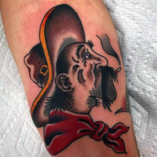 Tattoo of a rugged cowboy with a mustache, wearing a brown hat and red bandana, smoking a cigarette, done in bold traditional style.