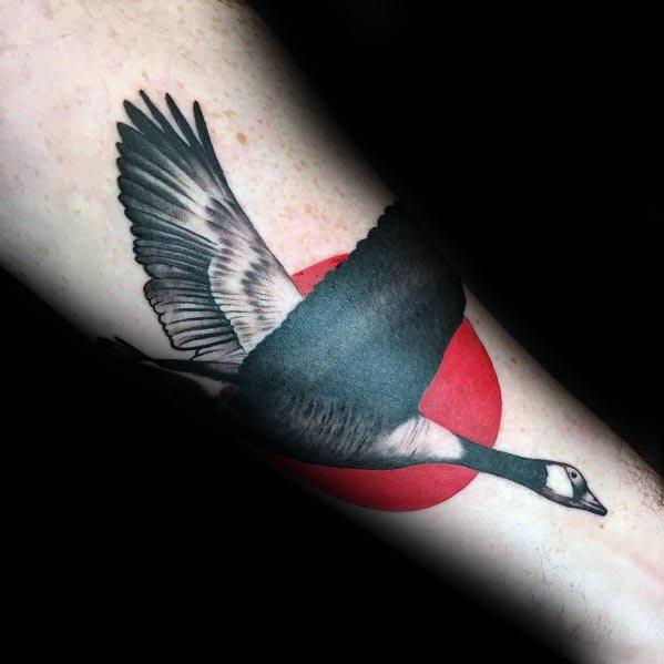 Realistic tattoo of a flying goose with detailed shading, set against a bold red circle, inked on the forearm for a striking contrast.