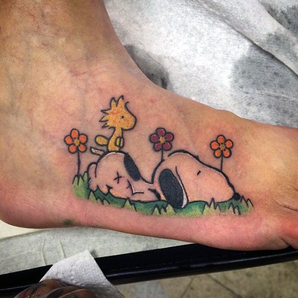 Colorful tattoo of Snoopy sleeping on grass with Woodstock standing on his back, surrounded by flowers, placed on a person's foot.