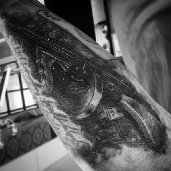 Realistic black and grey Tommy gun tattoo with intricate shading and depth, creating a bold and intense firearm design on the arm.
