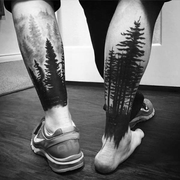 Black ink forest tattoos on both legs, featuring tall pine trees fading into mist, creating a realistic and immersive nature-inspired design.