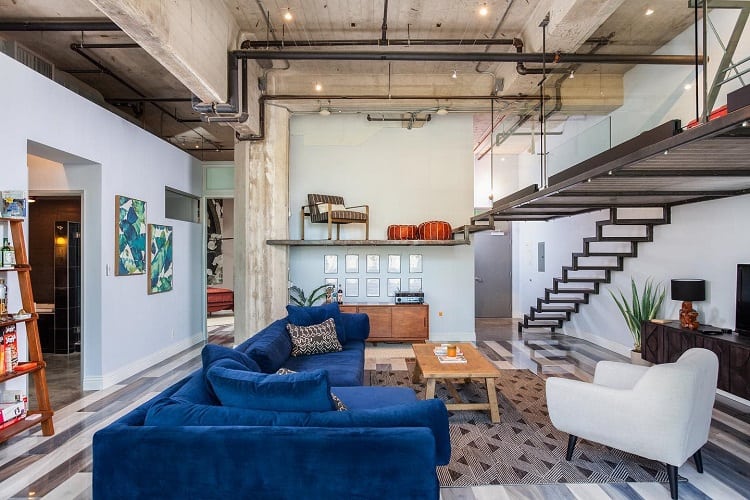 A stylish mancave loft with an unfinished concrete ceiling, industrial beams, and modern furniture, featuring a large blue sectional sofa, wooden coffee table, and open upper floor, creating a trendy and spacious urban vibe