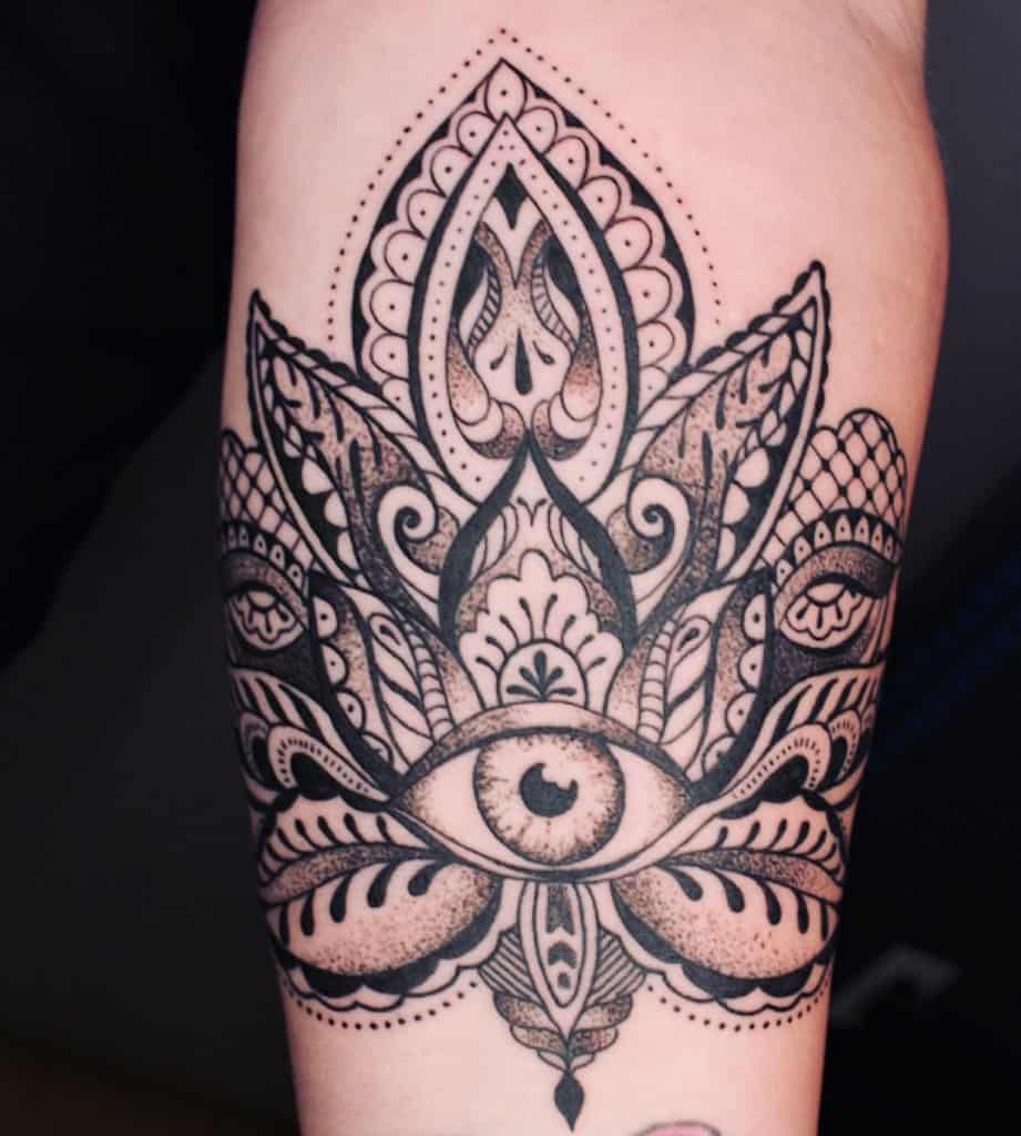 Tattoo of an all-seeing eye within an intricate ornamental lotus design, inked in black with dot shading on the forearm.