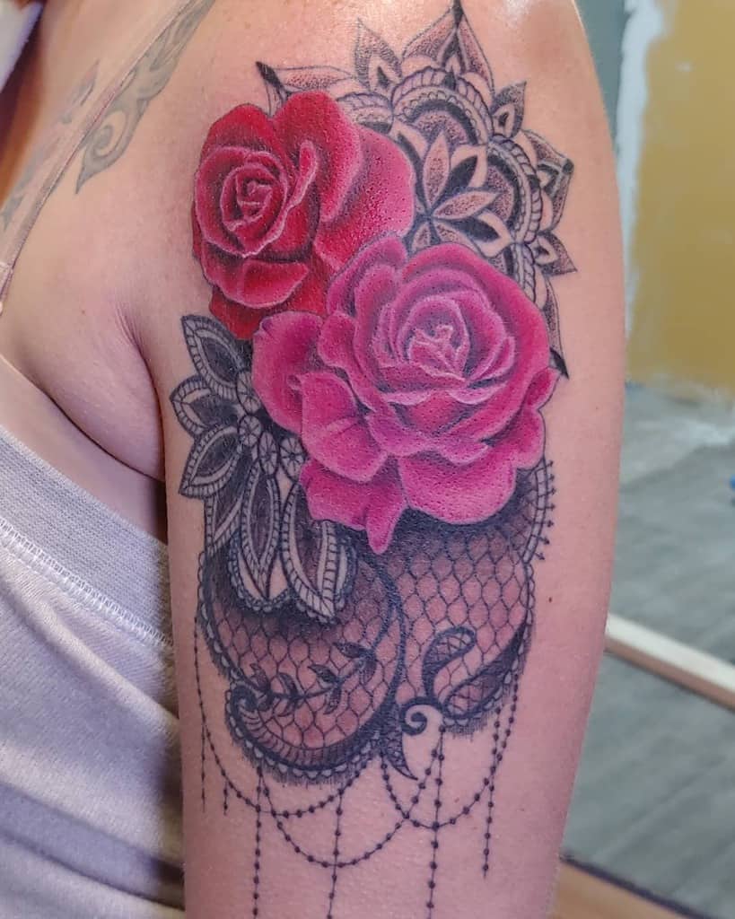Upper arm tattoo featuring vibrant red roses with intricate black lace, mandala details, and delicate beaded chain accents.