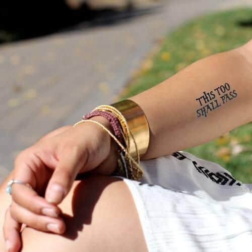 A person's arm with a "This Too Shall Pass" tattoo, wearing a gold bracelet and layered colorful bangles.