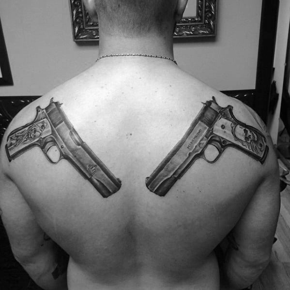 Black and gray back tattoo featuring two mirrored 1911 pistols on the shoulders, designed with intricate engravings and detailed shading for a bold look.