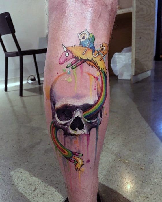 Colorful Adventure Time tattoo of Finn, Jake, and Lady Rainicorn flying over a realistic skull with dripping watercolor effects on the calf.