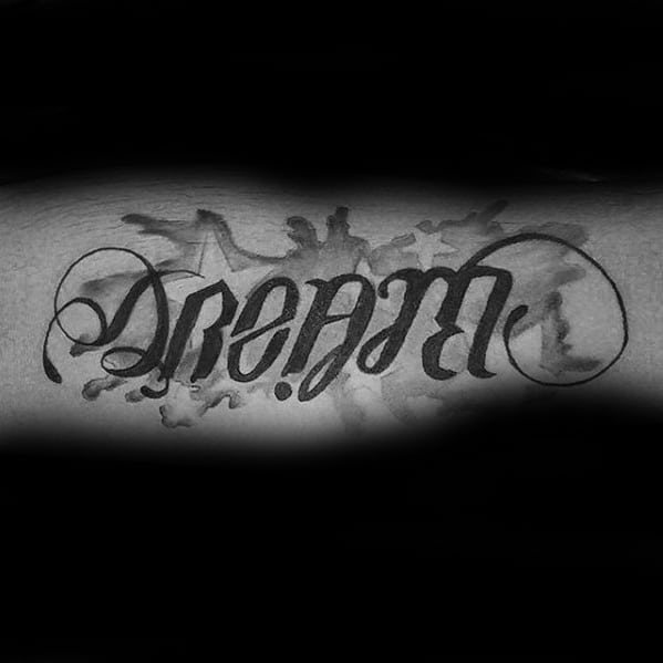 Forearm tattoo featuring the word “Dream” in an ambigram style, with artistic ink splashes and mirrored lettering for a bold visual effect.