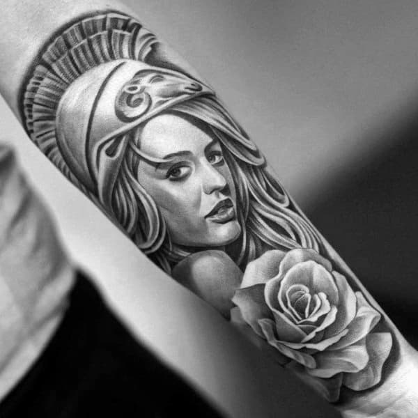Realistic Athena tattoo with soft facial features, detailed helmet, and blooming rose below in smooth black and grey shading on forearm
