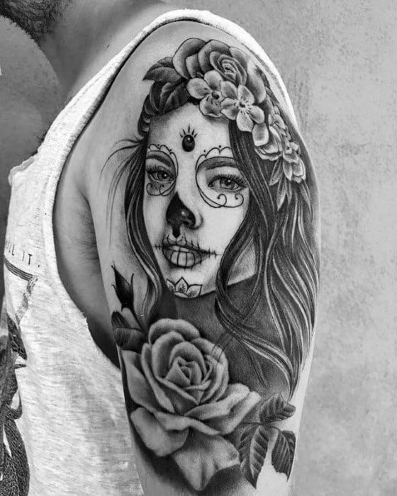 La Catrina tattoo on the upper arm showing a woman with sugar skull makeup, floral crown, and large roses in black and grey.