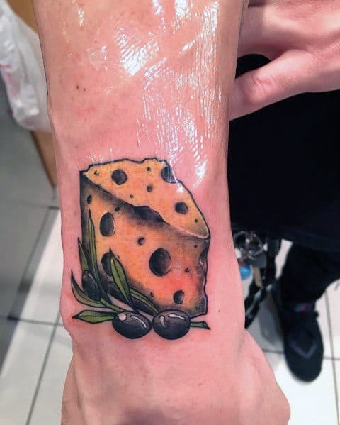 Colorful tattoo of a wedge of Swiss cheese with black olives and olive leaves, featuring bold outlines, smooth shading, and a realistic texture on the wrist.