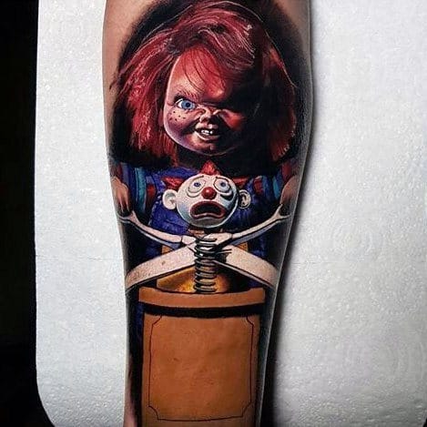 Realistic Chucky tattoo with vivid colors, featuring his sinister grin, glowing blue eyes, and a frightened jack-in-the-box clown for a chilling horror design.