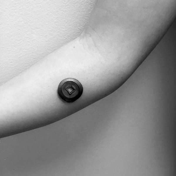 Minimalist tattoo of a small, circular coin with a square hole, placed on the inner arm, featuring clean shading for a subtle 3D effect.