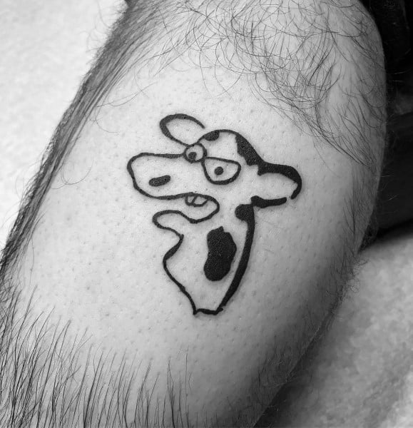 Small cartoon-style cow tattoo with bold black outlines, featuring a quirky expression and simplified spotted design.