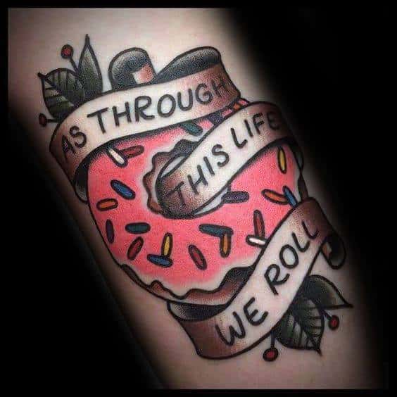 Tattoo of a bitten pink frosted donut with sprinkles, wrapped in a banner that reads as through this life we roll, with leaves and berries.