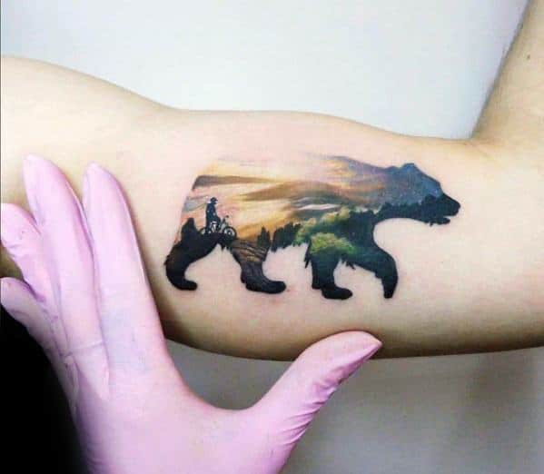 Tattoo of a bear silhouette filled with a scenic sunset landscape featuring a cyclist on a trail, symbolizing adventure and connection to nature.