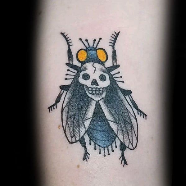 Creative fly tattoo with a skull face on its back, yellow eyes, and black-and-blue wings, blending creepy and cartoonish styles.