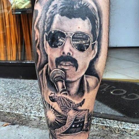 Detailed black and grey tattoo of Freddie Mercury with sunglasses, mic, and iconic stage pose, blending portrait and performance elements.