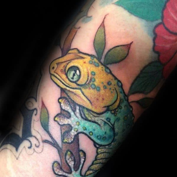 Colorful gecko tattoo with yellow and teal tones, perched on a branch among green leaves and a red flower background.